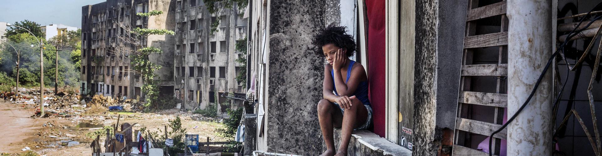 A colour photograph of a boy with a messy afro sitting on a window sill of slum-like apartment building. In the background two similarly abandoned looking buildings sit on the edge of a vacant lot strewn with litter.