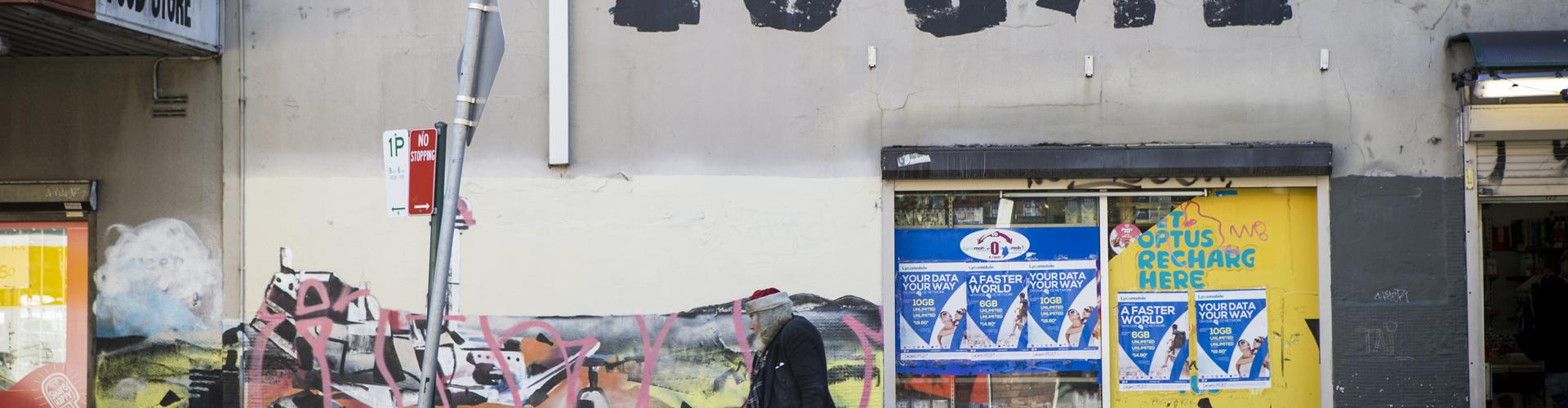 A colour photograph of a man wearing an old santa hat walking down a grafitied urban street. 