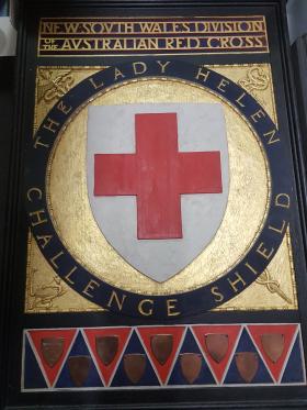 This beautiful woodcut shield, hand painted in gold and black with a red cross in the centre and decorated with brass plates, was designed by Professor L. Wilkinson of the University of Sydney and executed by Eirene Mort and Nora Weston.