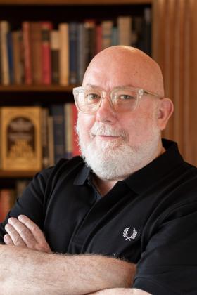 Photographic portrait of a bald, white bearded man, wearing clear framed glasses and smiling directly at the camea.