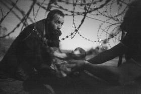 A black and white photograph showing a man passing a baby under a razor wire fence to a person waiting on the other side.