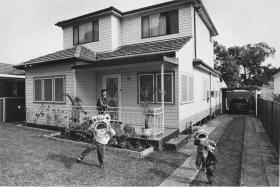 Three children wearing Chinese dragon paper heads play in a suburban front yard, their father watching on from the front verandah.