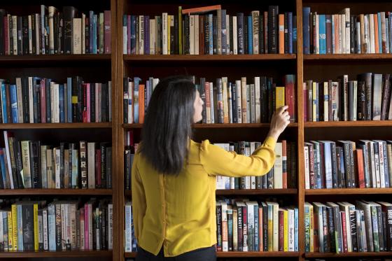 A woman in a yellow shirt with dark hair pulls a book from a large bookshelf