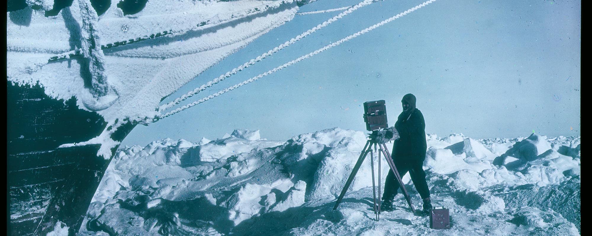 A man operates a large 20th century camera amid a snowy tundra, in front of a docked ship.