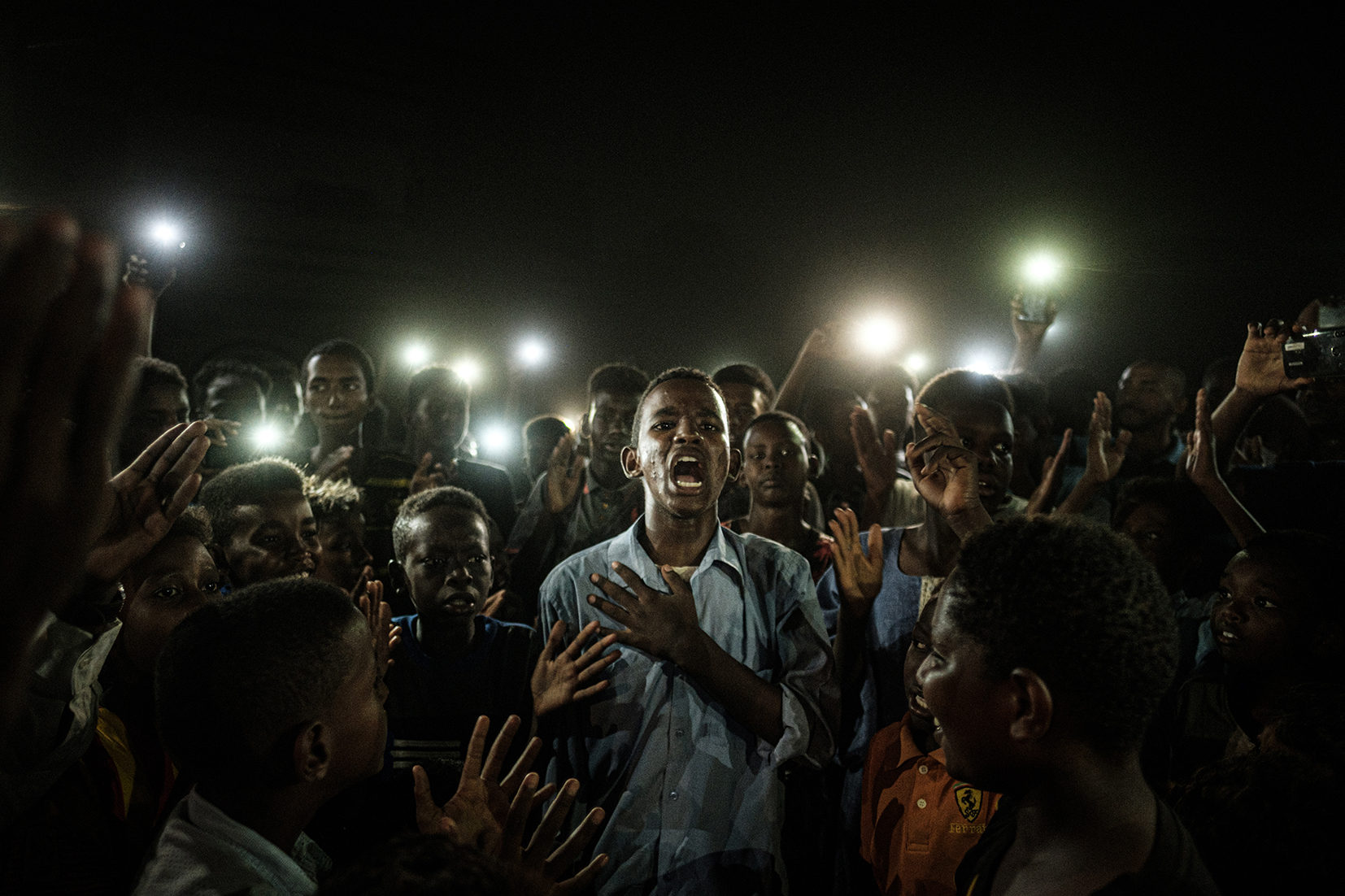 A young Sudanese man stand with his hand on his heart, mid sentence. He is surrounded by a crowd holding up their mobile phones to illuminate him. 