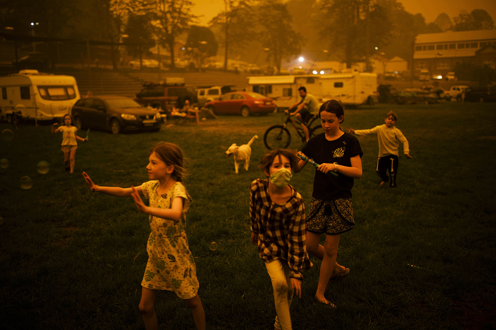 Photograph featuring children playing on a sports field. One wears a face mask. The sky and light are tinged orange with smoke. 