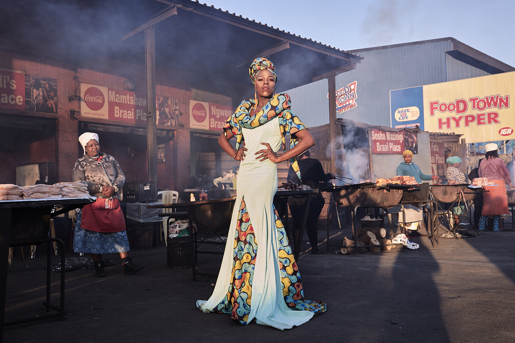 Photograph of  man standing in front of BBQ food vendors in an African market - he stands tall gazing at the camera with hands on hips wearing a long colourful dress, head scarf and makeup.