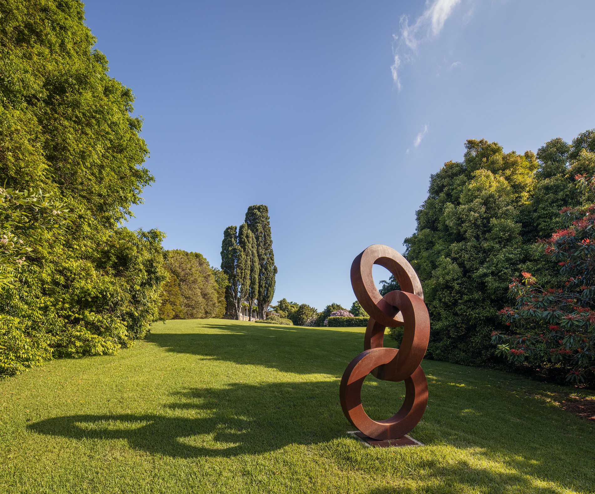 A photograph of a large metal sculpture of interlocking circles stacked vertically. The sculpture sit in a garden, with a house in the background.