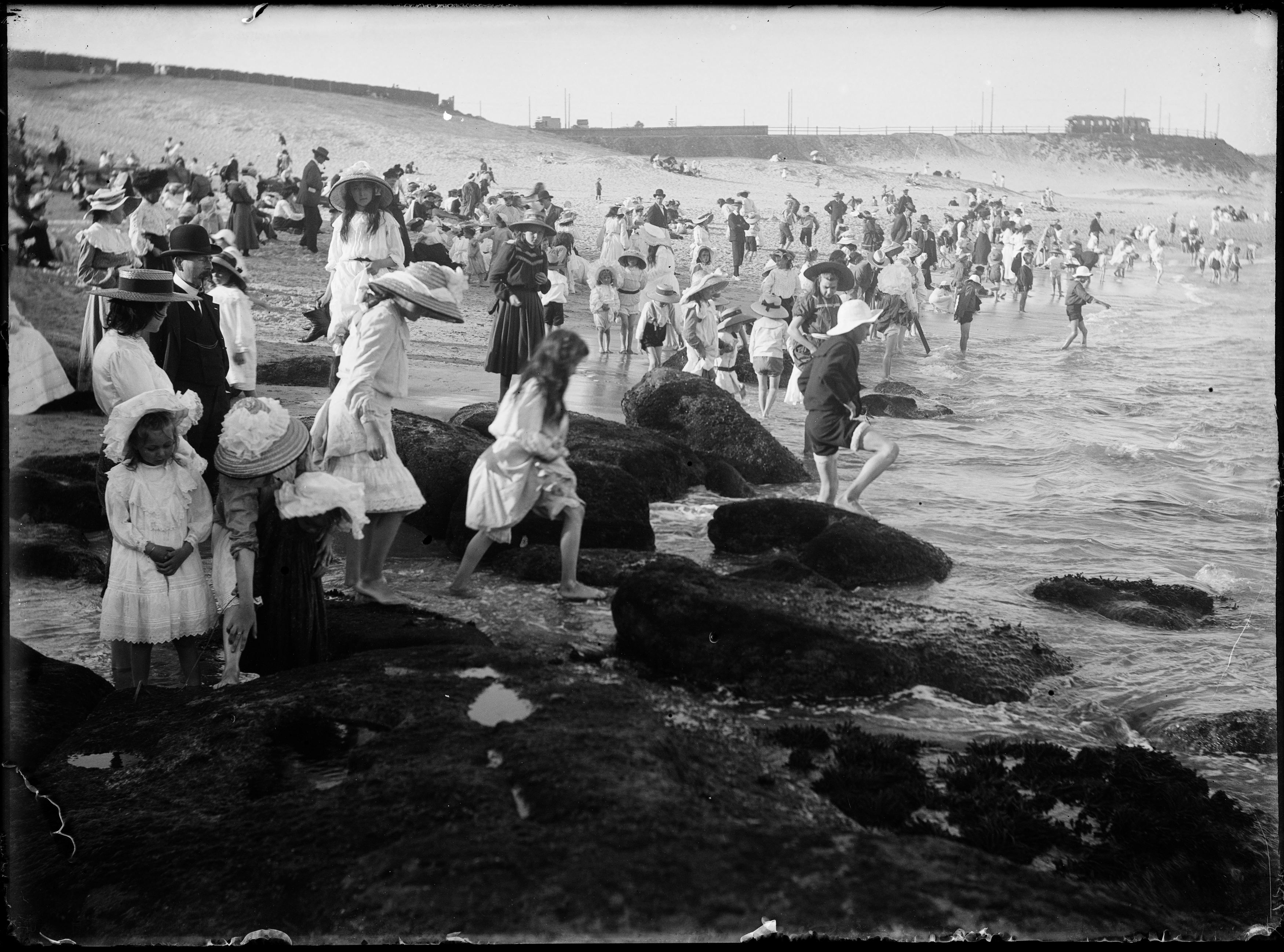 A black and white photograph of a crowded beach. Most people are fully clothed in 19th Century dress. 