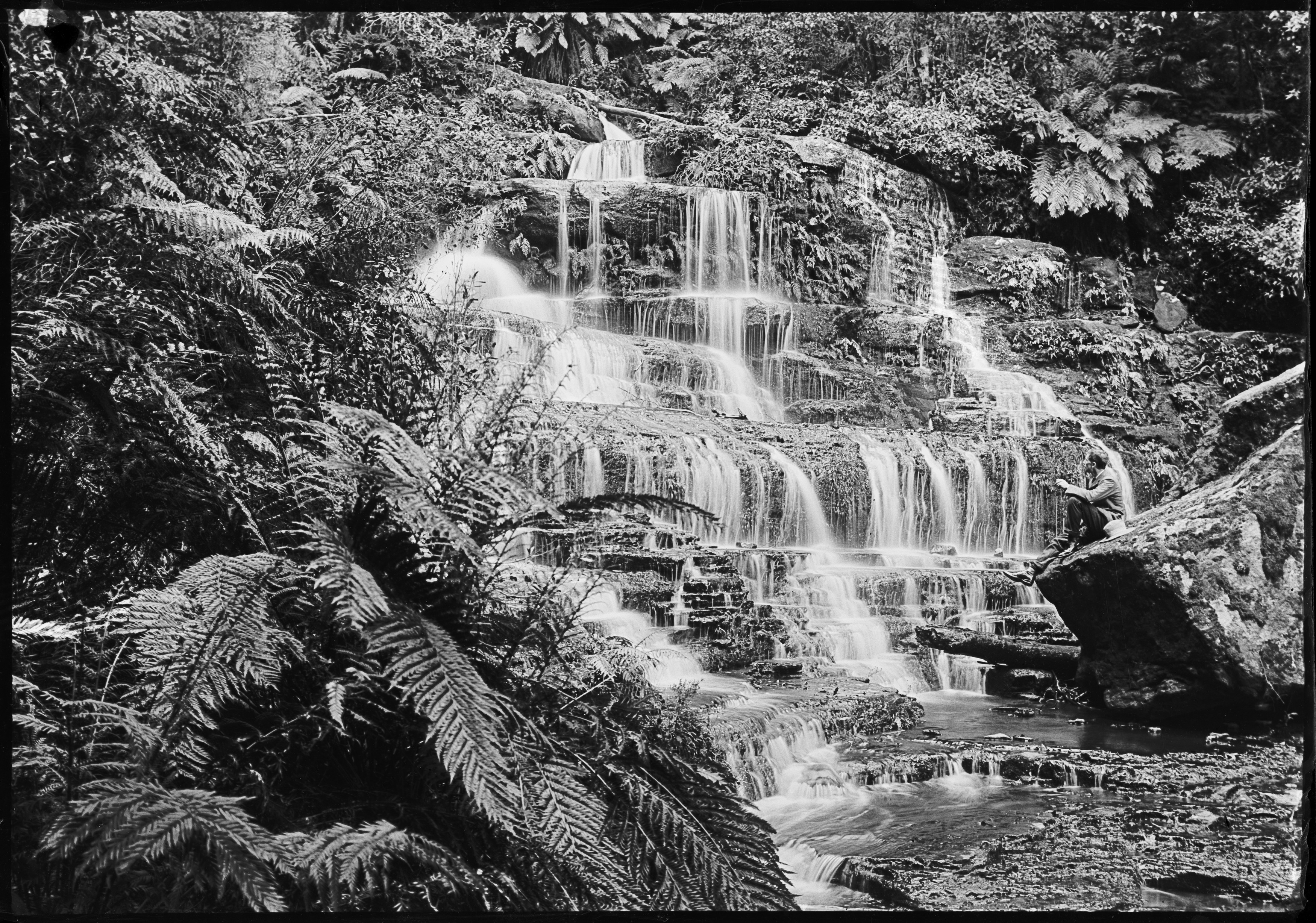 Black and white photograph of a cascading waterfall. A man sits on a rock in the foreground.