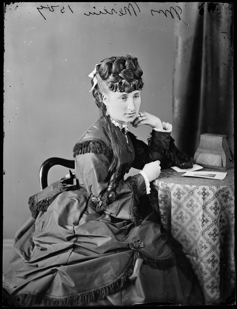 An old black and white photo of a woman with an elaborate hairstyle, in a contemplative pose sitting at a small round table.