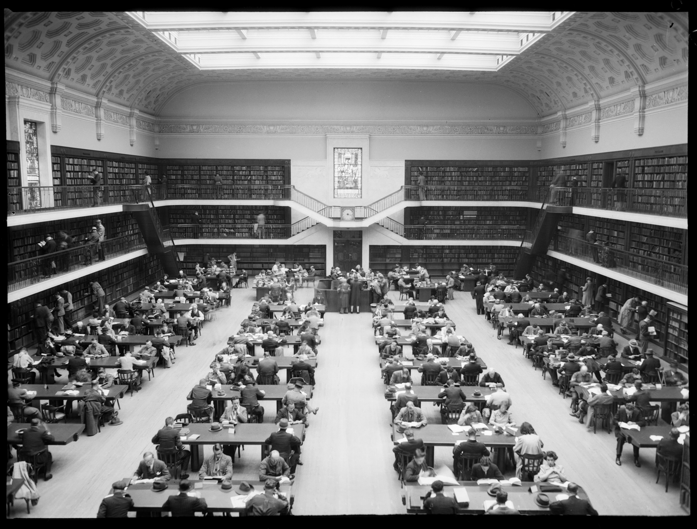 Large room in Library with glass ceiling and books shelves around the circumference of the room. People at desks reading. 