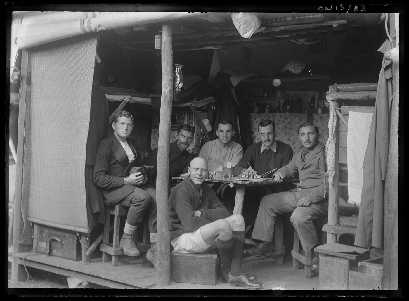 Black and white photograph of men sitting around a table playing chess, in a make shift shelter.