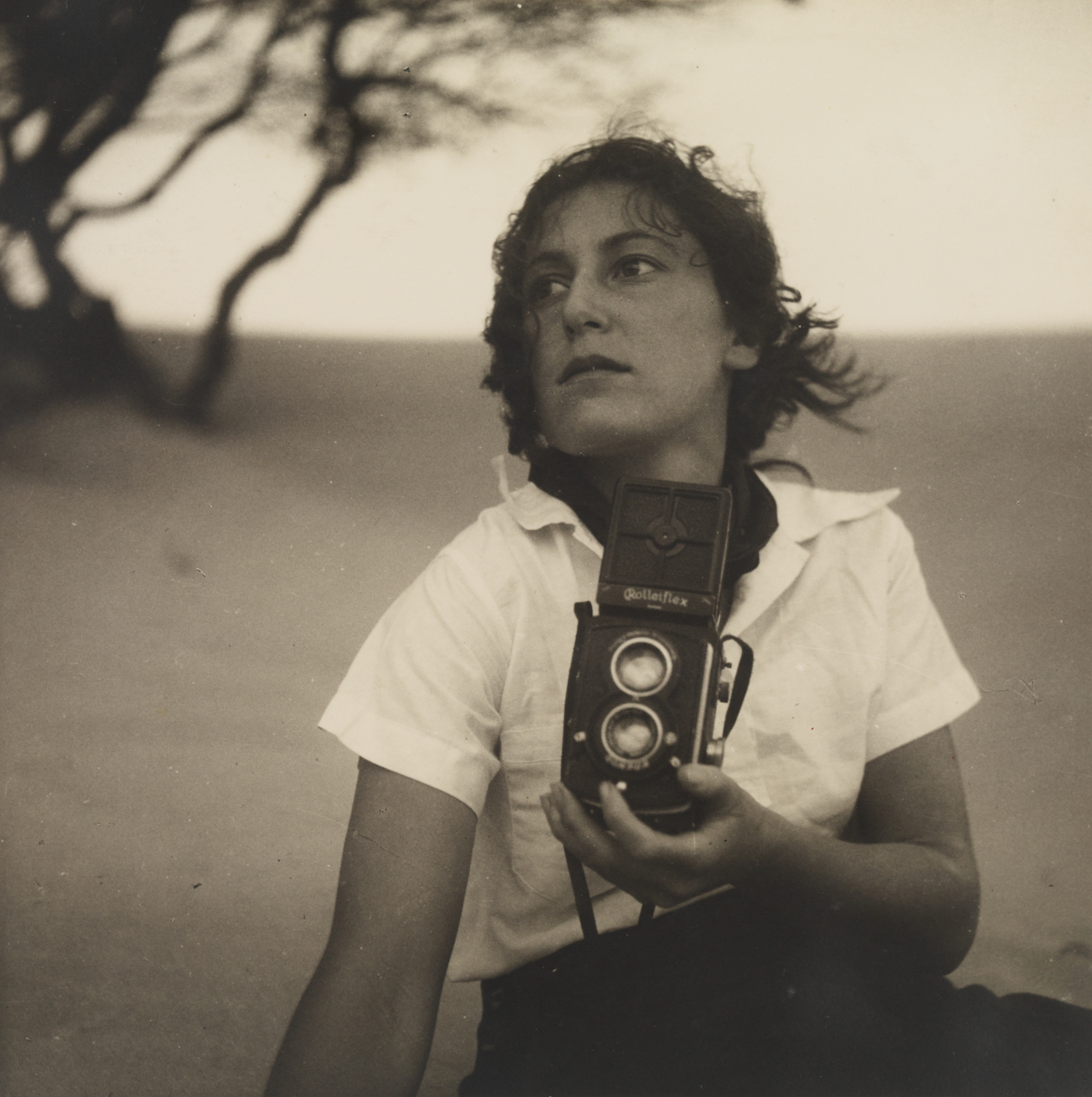  A sepia photographic portrait of a woman sitting on a beach gazing off to the size, holding an old camera up in front of her
