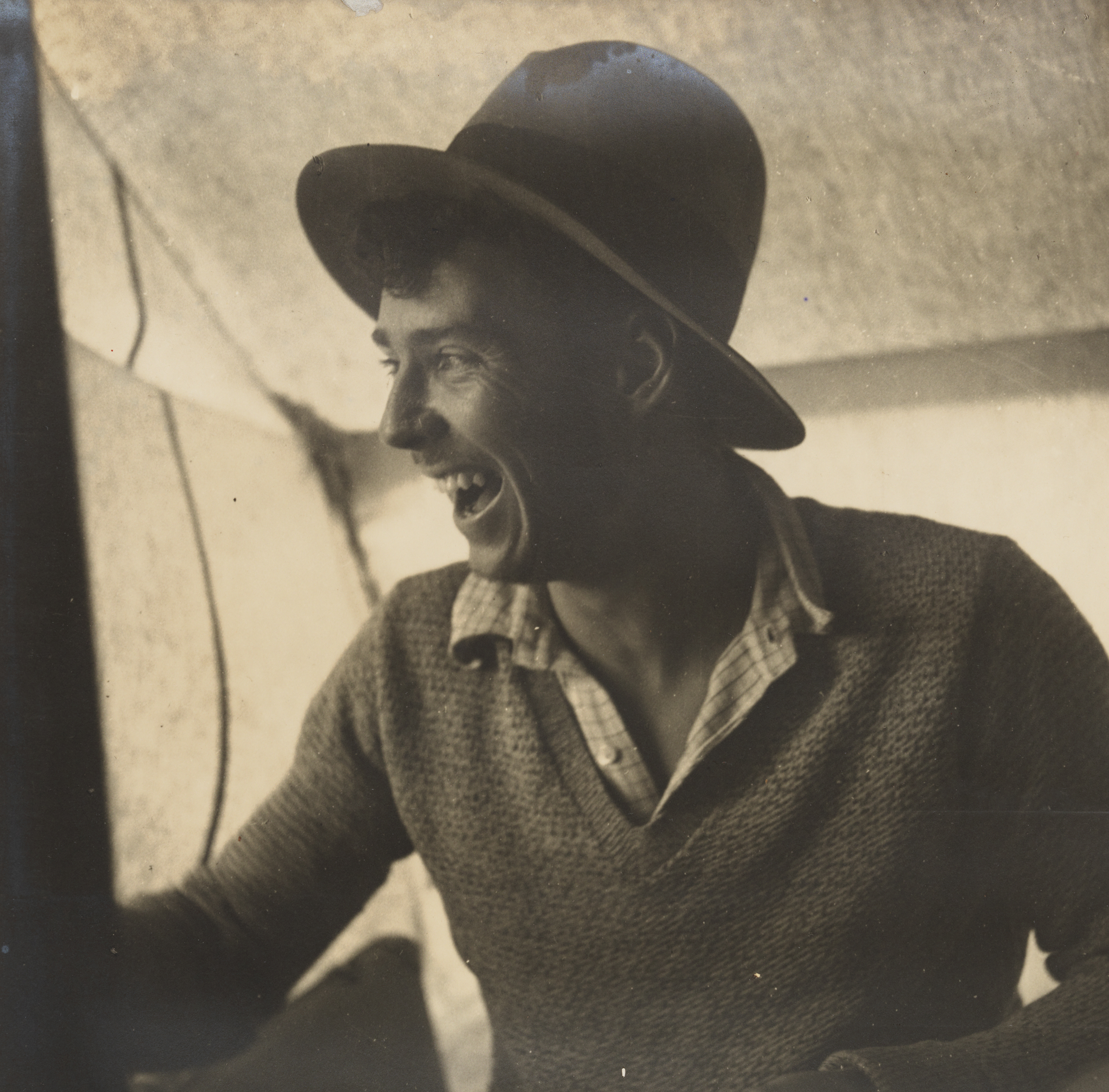 A sepia photograph of a man wearing a hat, checked shirt and wollen jumper sitting inside a canvas structure, looking off to the side laughing.