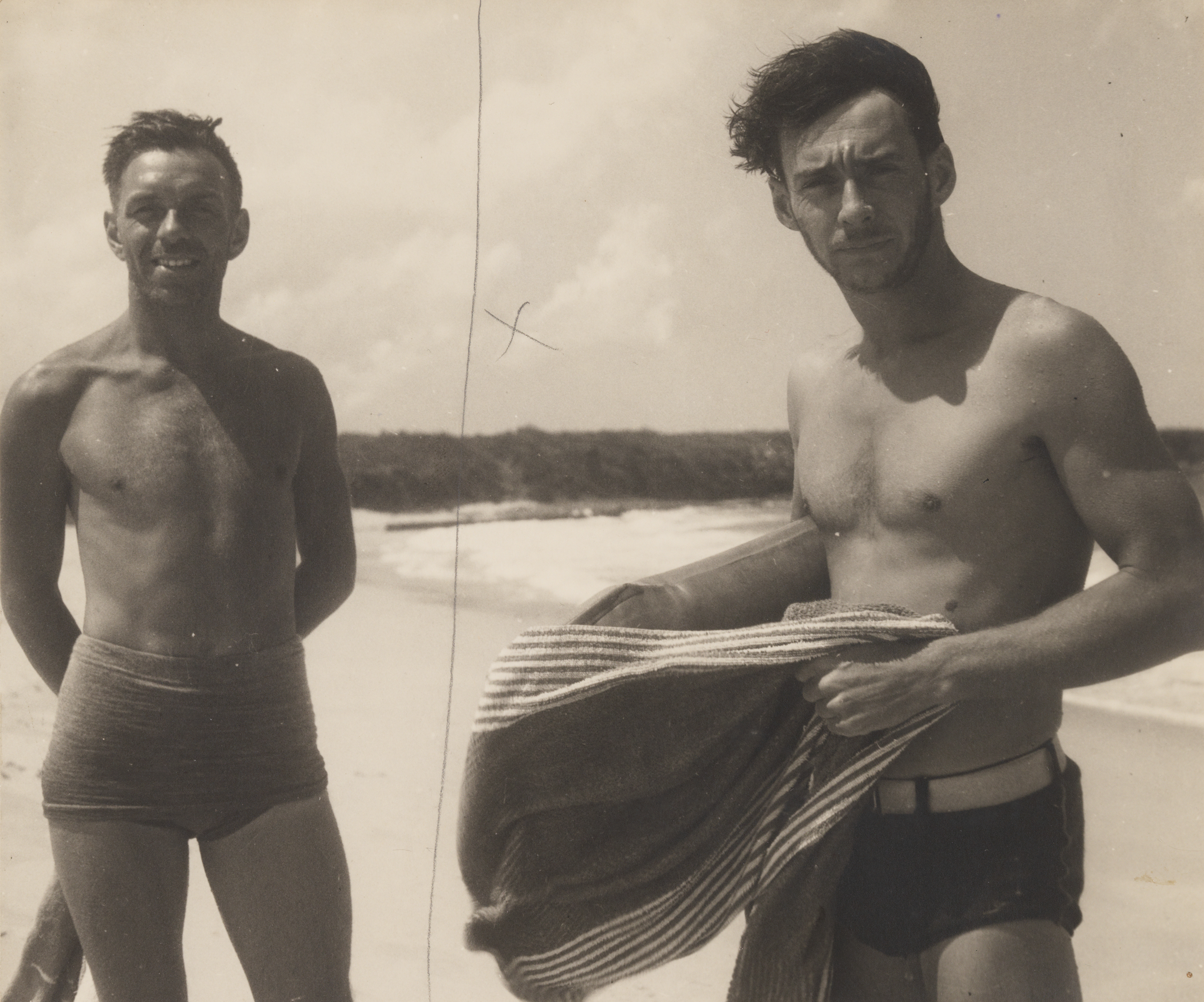 A sepia photograph of two men standing side by side on a beach, looking at the camera smiling - they wear 1930s style bathing shorts and hold towels