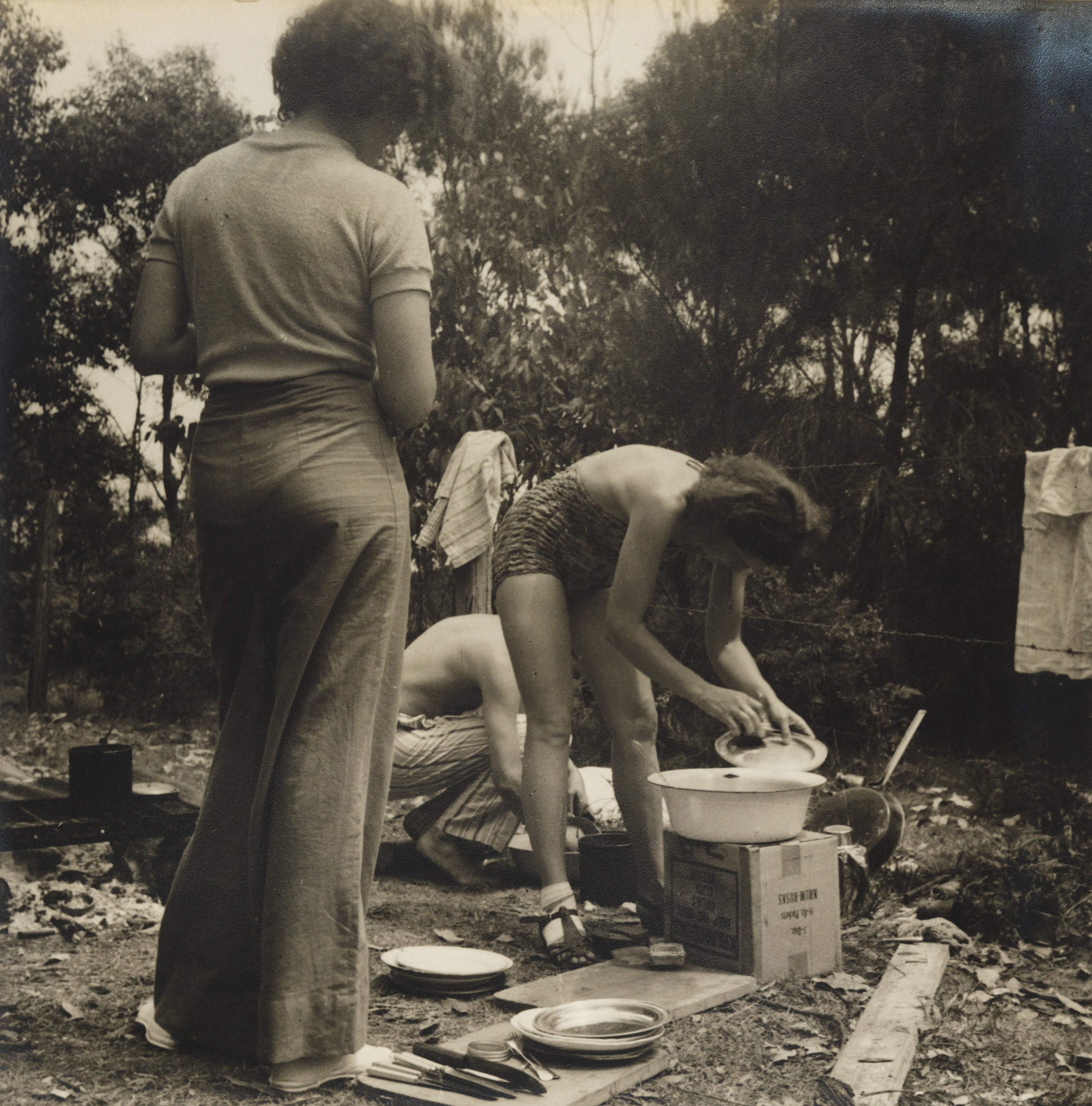A sepia photograph of a woman washing up tin plates at a camp site - next to the camp hot plate. 