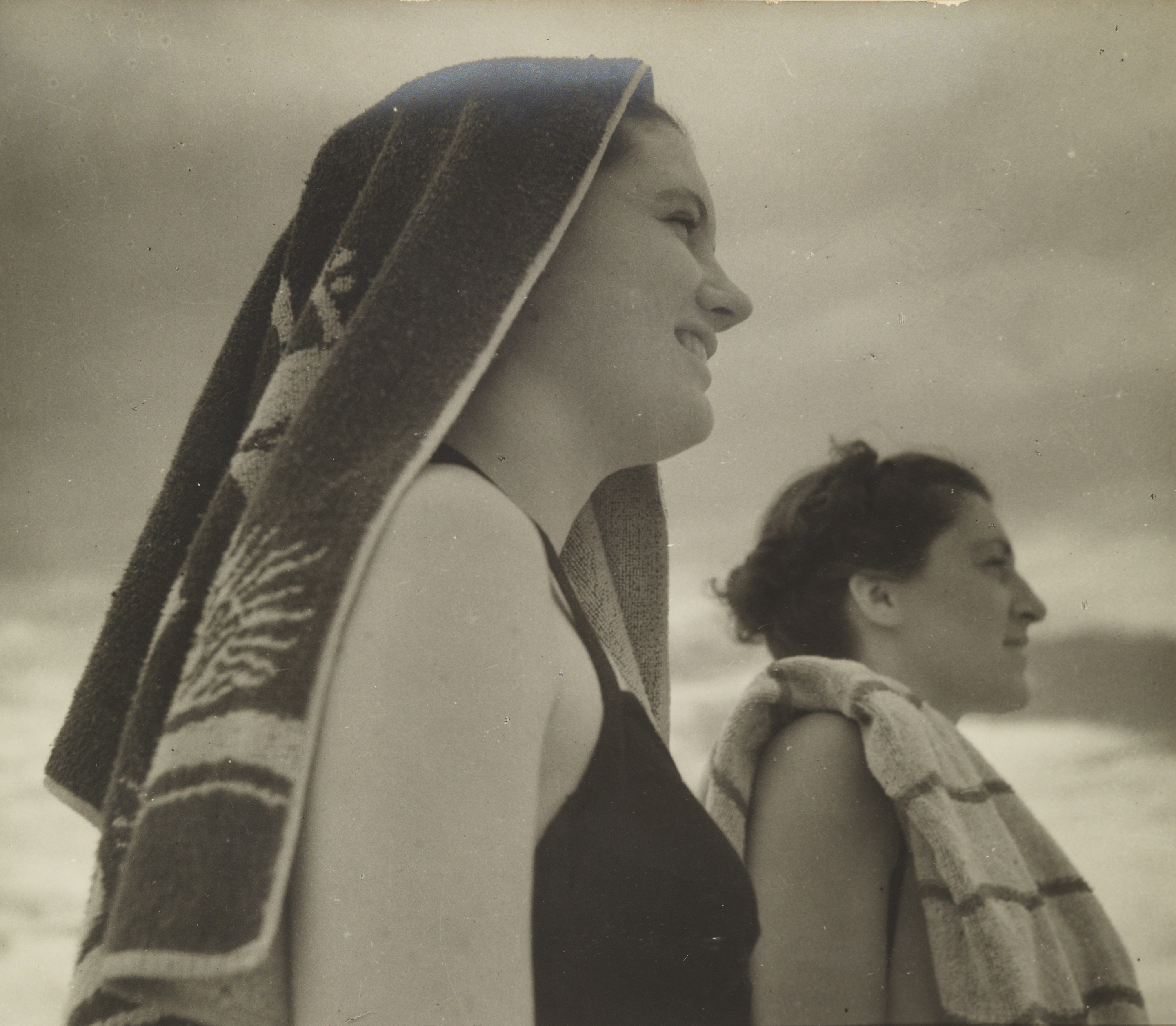 A sepia photograph of two women standing side by side in profile. The woman in the foreground has beach towel draped over her head.