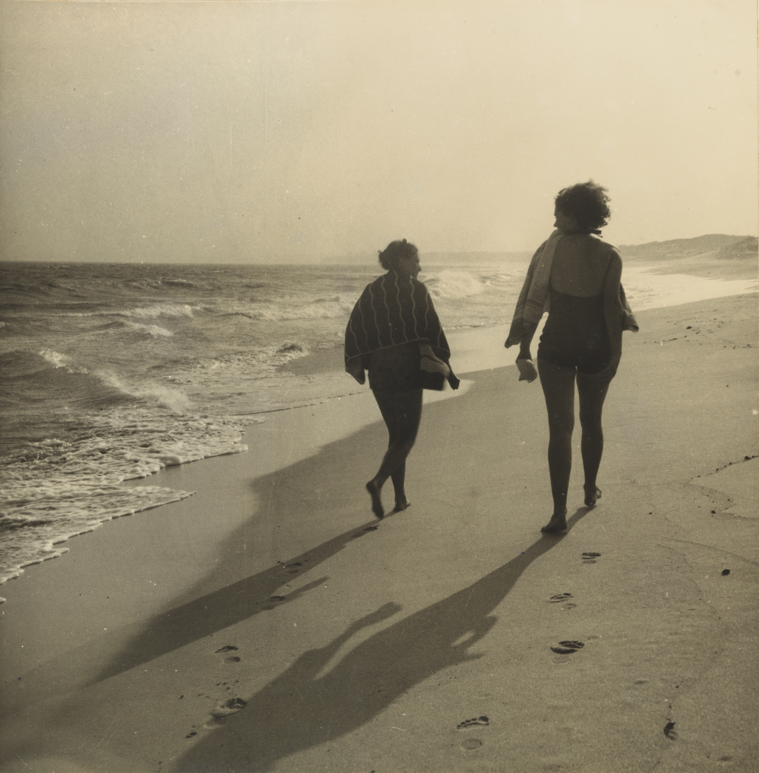 A sepia photograph of two women walking along the shoreline of a beach.