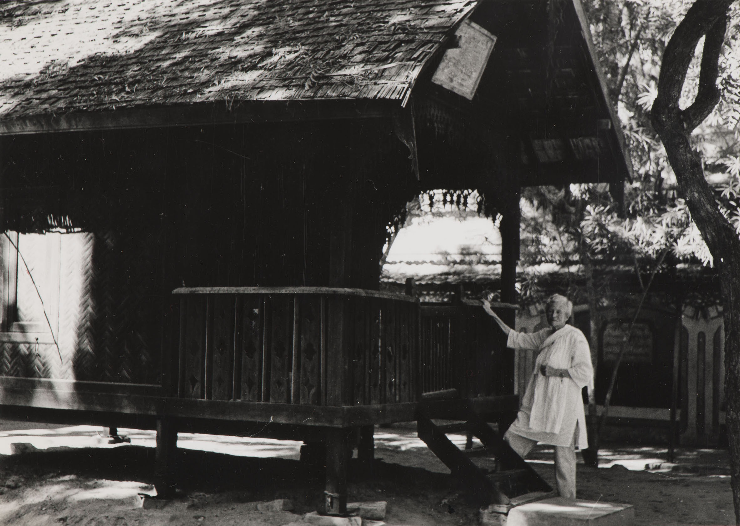 A black and white photograph of an older woman standing next to a hut, resting her hand on the banister.