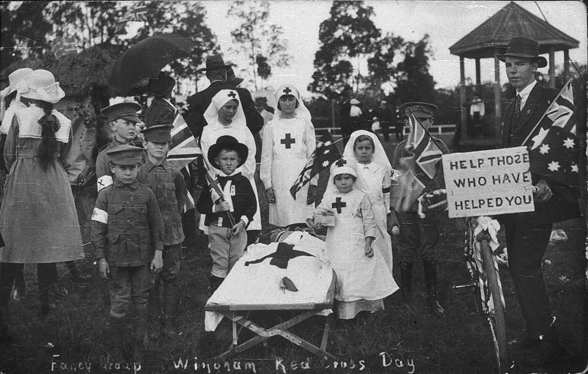 Black and white photograph of children dressed up in nurses and medics posing for the camera. A mock stretcher stands in the middle of the group. 