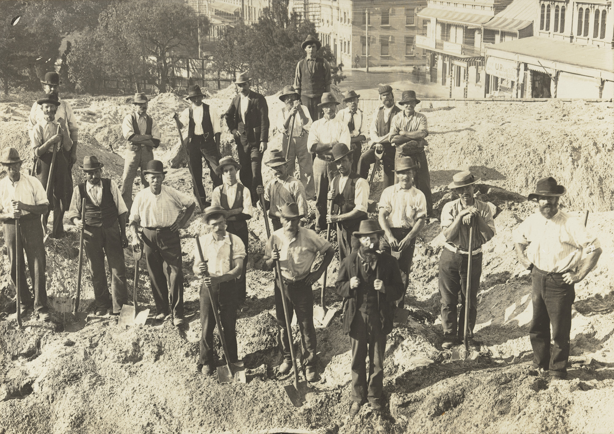 A sepia photograph of a group of workers, standing facing the camera, shovels in hand - they wear trousers, button up shirts and suspenders. 