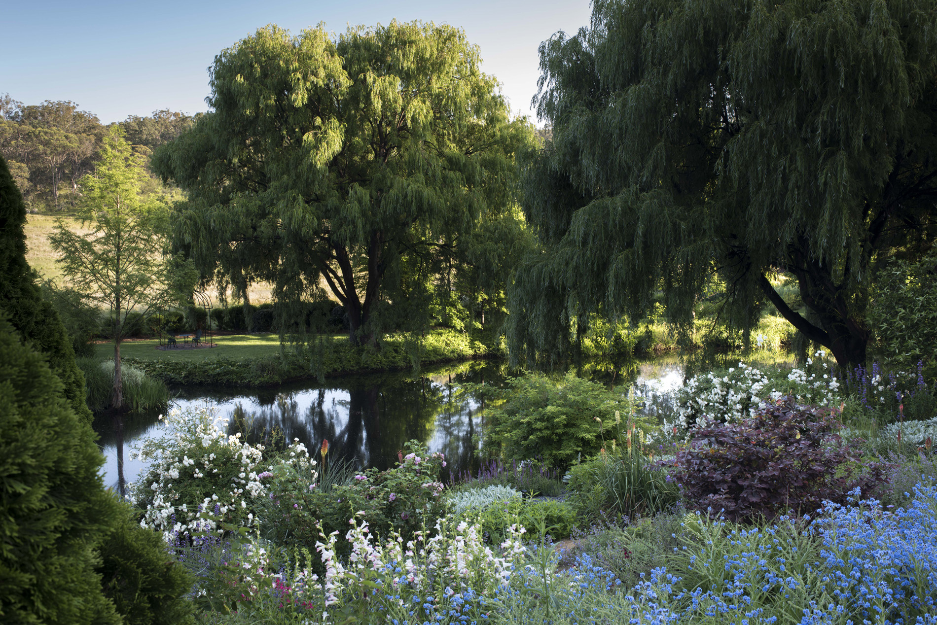 A photograph of a garden. Flowers and a lake sit in the foreground, with lush green grass backing onto a dry paddock in the background.