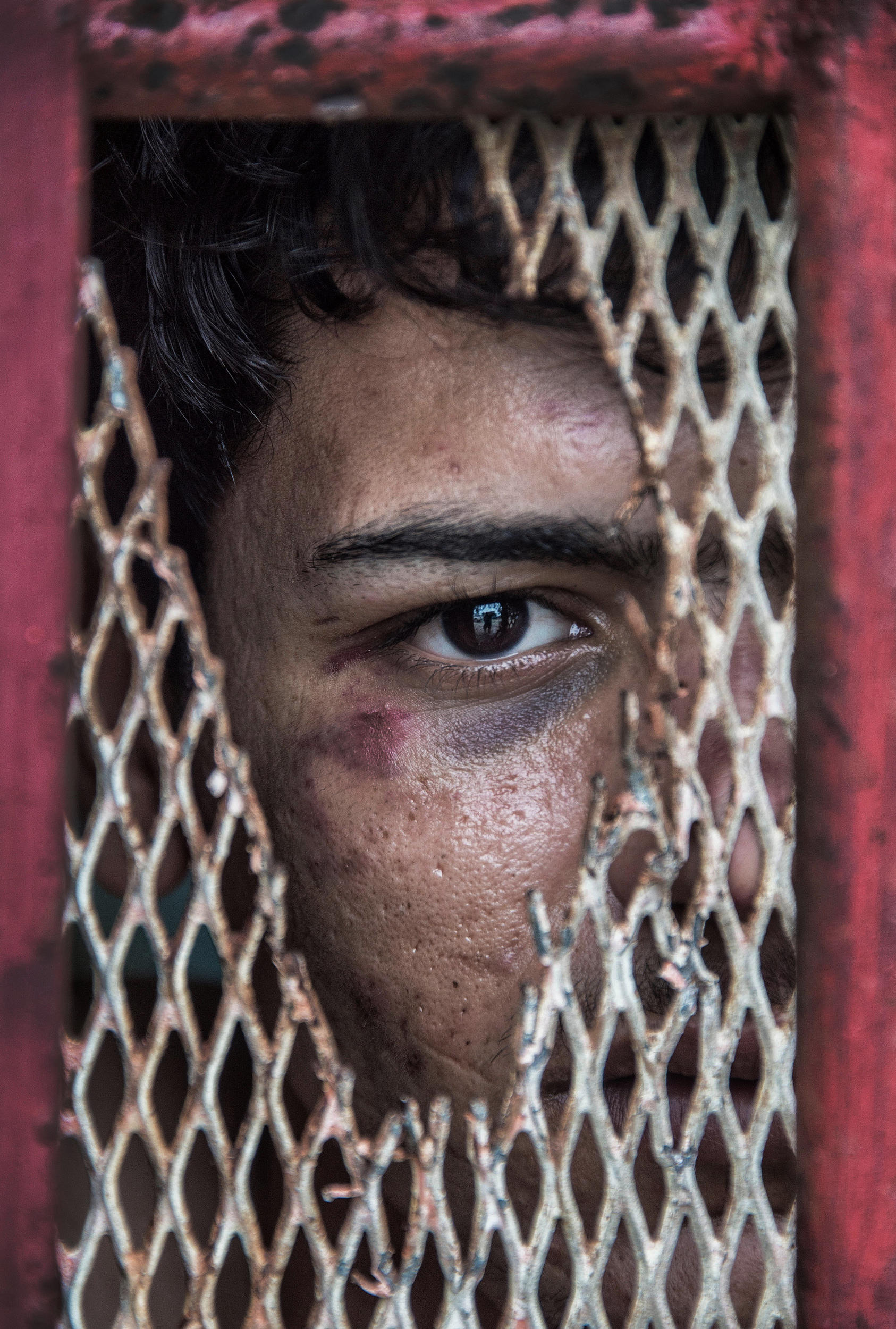 A close up colour photograph of a boy looking through the hole in a broken metal grate. 