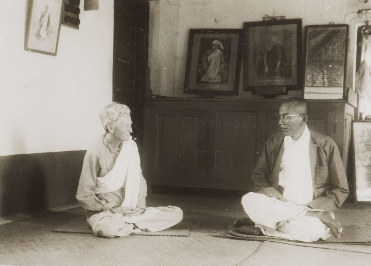 An older woman and an older man wearing Buddhist monk clothes sit cross-legged next to each other.