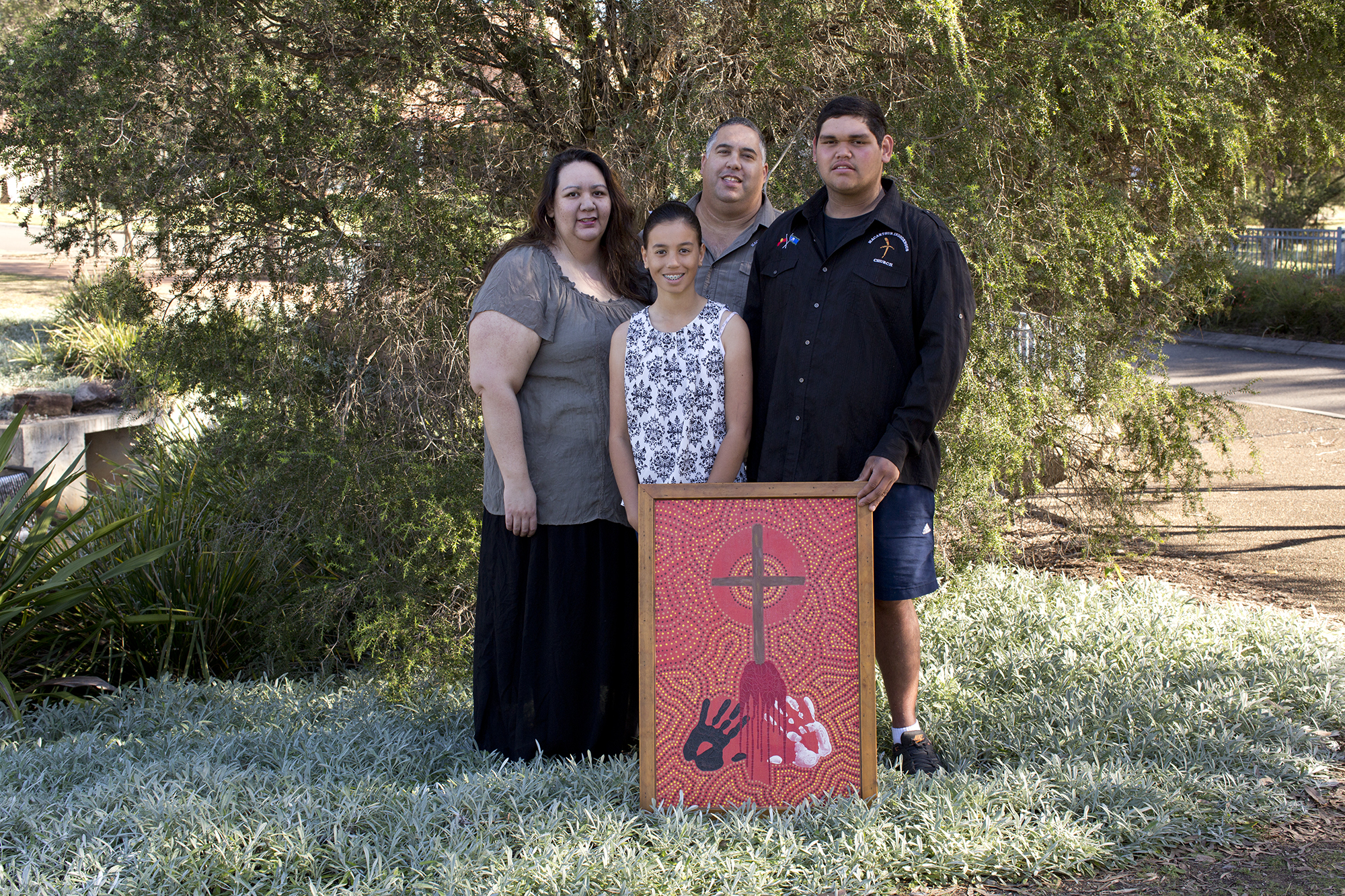 A family of four poses for the camera in front of a tree with an Aboriginal artwork.