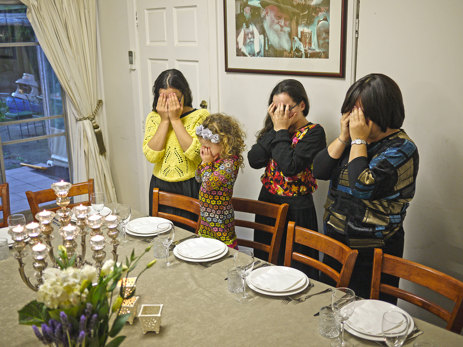 Three girls and a woman stand in front of settings at a dining table with their hands covering their eyes.