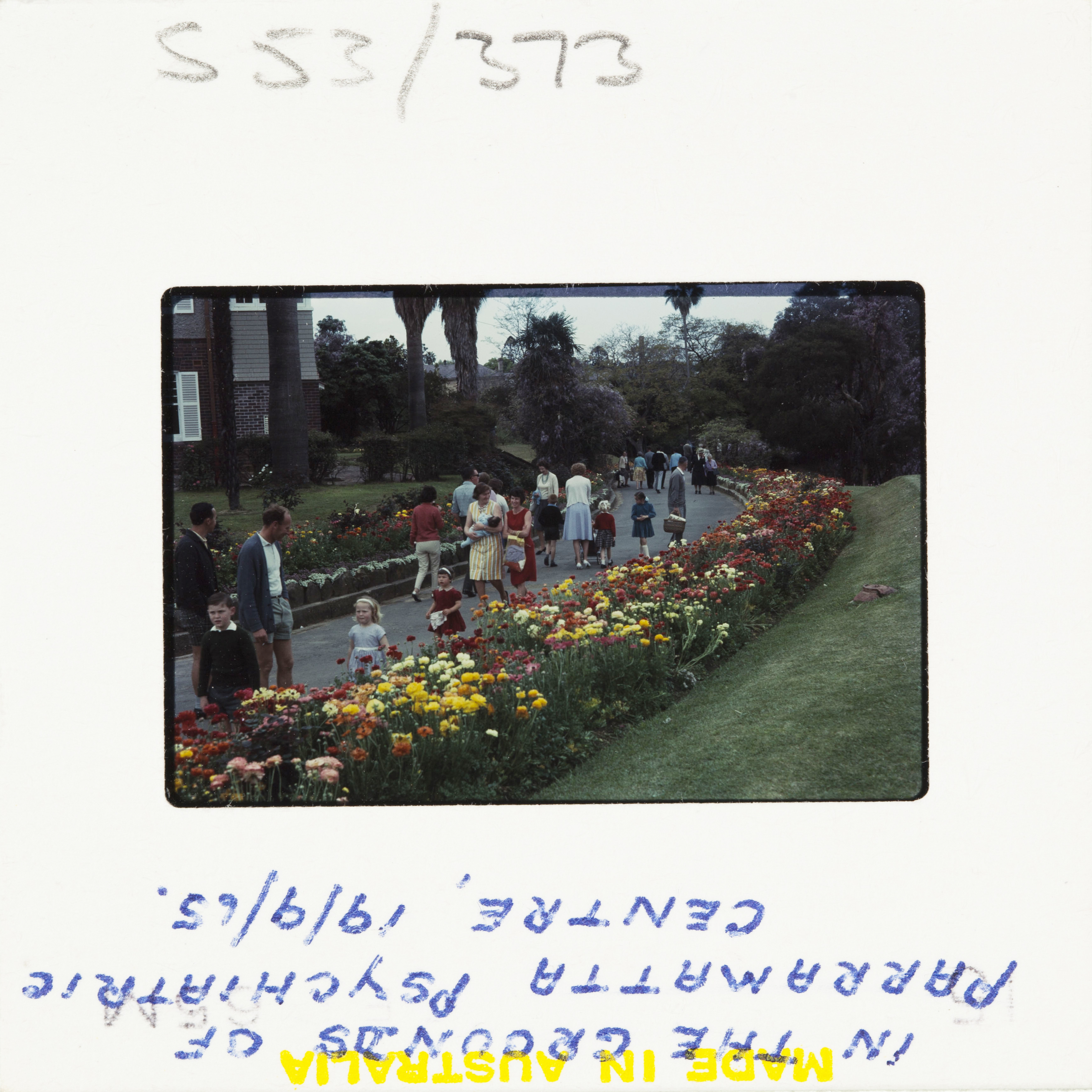 A 60s Kodak colour slide showing people walking down a path with flower beds on either side. 