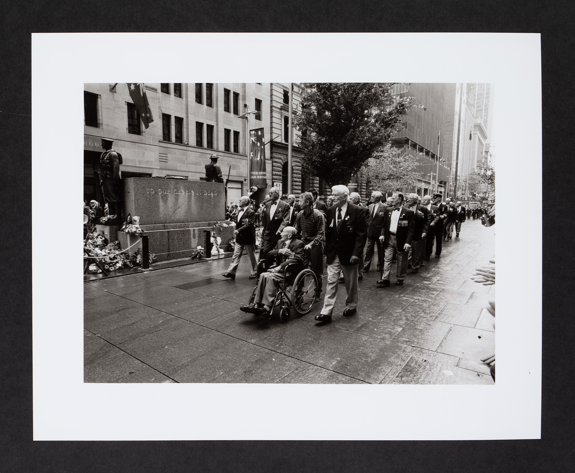 A marching contingent of veterans, one in a wheelchair, past a war memorial.