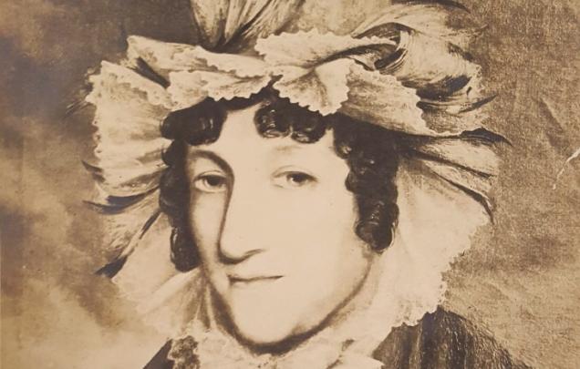 A portrait of a woman wearing a very ruffled bonnet, ruffled blouse and coat.