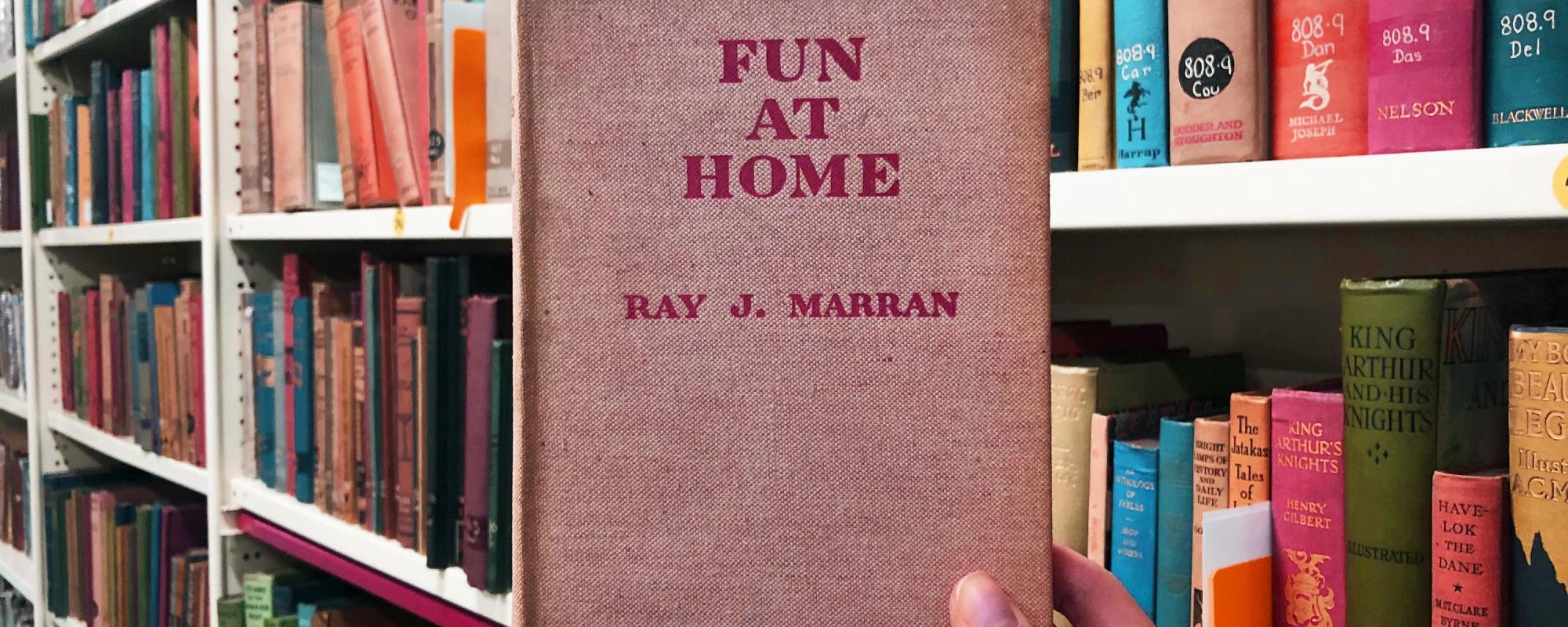 Hand holding up book titled 'Fun at Home' with book shelves in the background