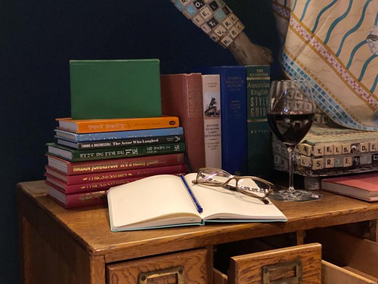 A wooden bookshelf has piles of books on top of it, and an open notebook with a pencil and a pair of glasses resting on top. Next to it is a glass of red wine.