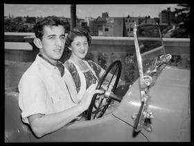 A black and white photo of a couple sitting in an open top 1950s car.
