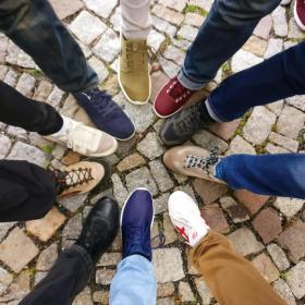 A group of friends standing on the cobblestones ground making foot circle with their legs and different shoes.
