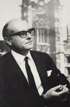 Image of a man in a suit, wearing black, square-rimmed glasses and holding a cigarette.