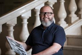 Matt Devine sits on some marble stairs holding a print of a black and white photograph. He has a grey beard and black glasses, and is wearing a navy shirt with white spots. He smiles at the camera.