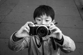 A black and white photo of a small boy holding a camera up in front of his face to take a photo - he is standing on concrete paving and wearing up a zip up parka.