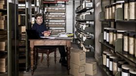 Young man sits at a wooden desk, surrounded by shelves of boxes and jars.