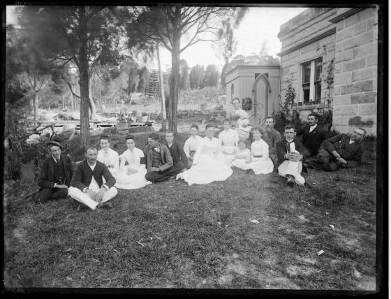 Black and white photograph of family sitting on the grass next to a sandstone homestead. 