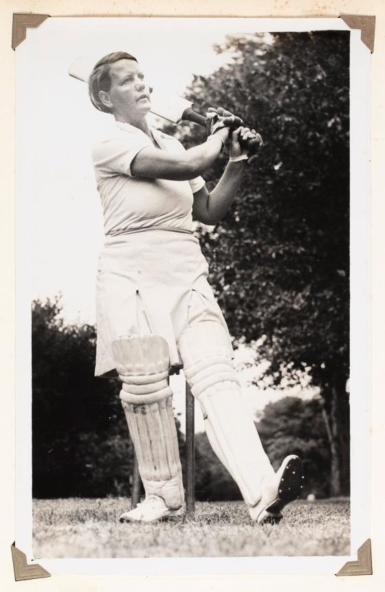 A woman in cricket uniform, swinging a cricket bat.
