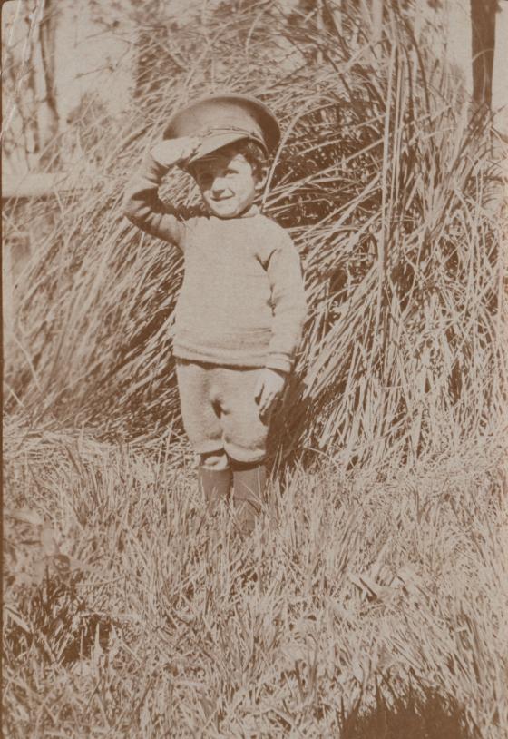 A sepia photograph of a boy wearing an oversized army cap, standing and saluting.