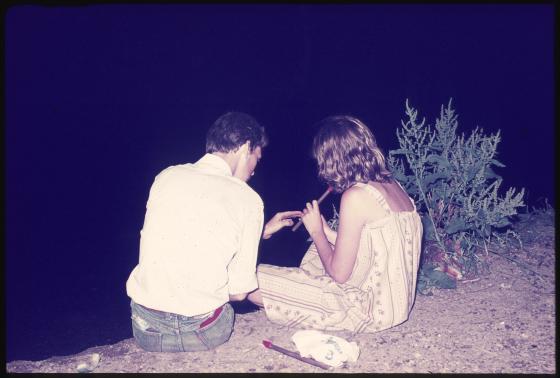 A man shows a woman how to play a pennywhistle while sitting on the bank of a river at night