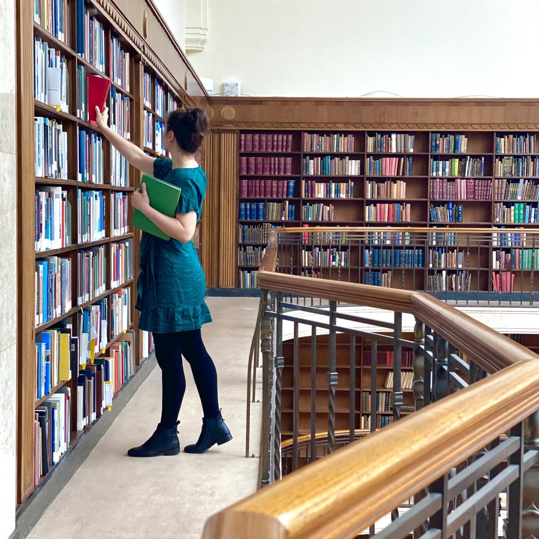 Woman stands in Mitchell Library Reading Room, pulling a book off bookshelf.